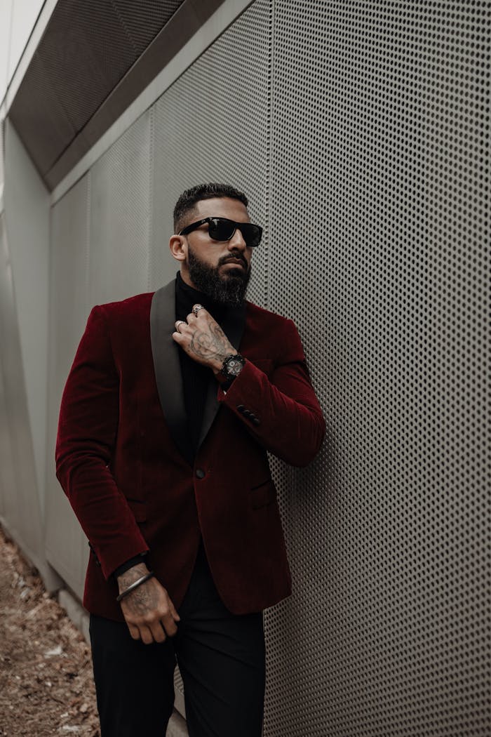 Confident man in a red velvet blazer and sunglasses leaning on a modern metal wall.