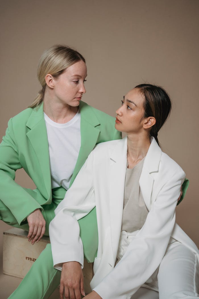 Two women in pastel blazers share a moment in a minimalist studio portrait.